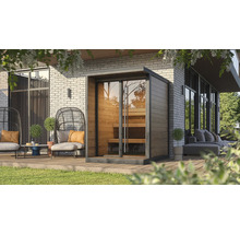 Sauna extérieur moderne en bois avec façade vitrée et bancs intérieurs sur une terrasse en bois avec mobilier de jardin devant un mur en briques.