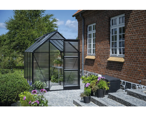 Gewächshaus mit geöffneter Tür in einem Garten neben einem Backsteingebäude.