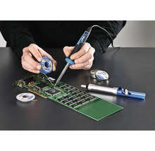 Une personne soude une carte de circuit imprimé avec un fer à souder, de l'étain à souder et une pompe à dessouder.