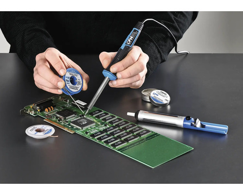 Une personne soude une carte de circuit imprimé avec un fer à souder, de l'étain à souder et une pompe à dessouder.