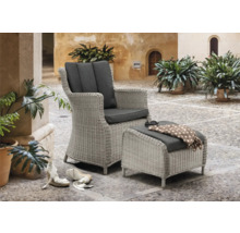 Geflochtener Sessel mit Fusshocker und grauen Polstern auf einer gepflasterten Terrasse.