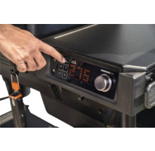 Main utilisant le panneau de commande numérique d'un barbecue Traeger avec affichage de la température, bouton de réglage et Traeger Logo.