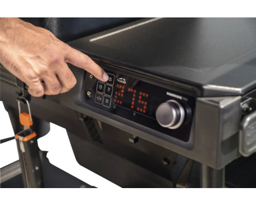 Main utilisant le panneau de commande numérique d'un barbecue Traeger avec affichage de la température, bouton de réglage et Traeger Logo.