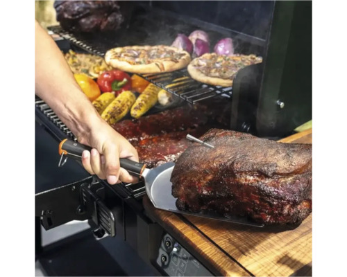 Scène de barbecue avec de la viande, des pizzas, des épis de maïs et des légumes préparés sur un gril. Une personne tient une spatule.
