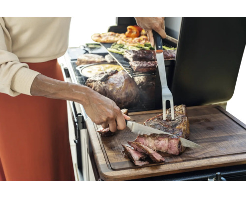 Une personne coupe de la viande grillée sur une planche à découper à côté d'un barbecue contenant d'autres viandes et légumes.