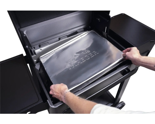Mains plaçant une protection de plateau d égouttement en aluminium Traeger dans un barbecue à pellets Traeger noir.