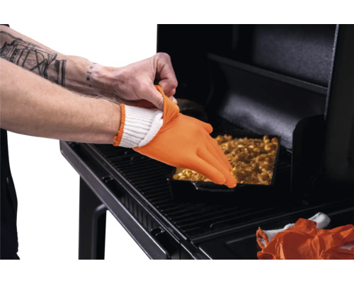 Utilisation d'un gant de barbecue en silicone orange résistant à la chaleur sur un gant en tissu blanc devant un barbecue avec un plat.