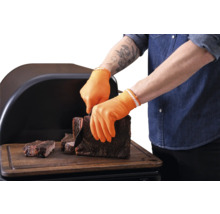 Personne avec gants de barbecue orange résistants à la chaleur coupant de la viande grillée sur une planche en bois devant un fumoir.