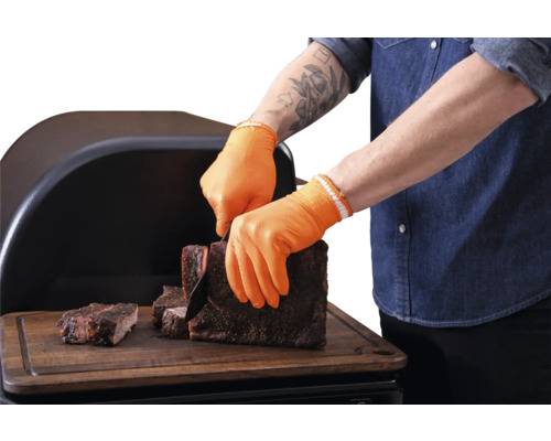 Personne avec gants de barbecue orange résistants à la chaleur coupant de la viande grillée sur une planche en bois devant un fumoir.