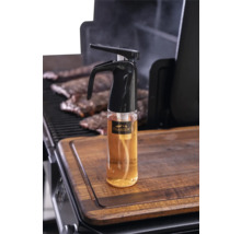 Vaporisateur d'huile Traeger avec Traeger Logo sur une planche à découper en bois à côté d'un barbecue avec des travers de porc.