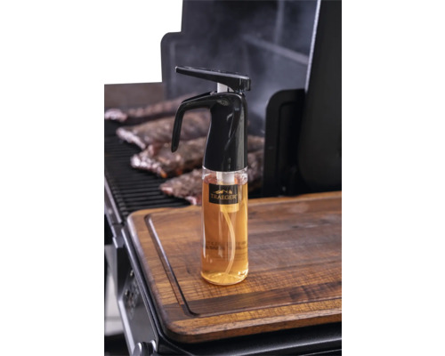 Vaporisateur d'huile Traeger avec Traeger Logo sur une planche à découper en bois à côté d'un barbecue avec des travers de porc.
