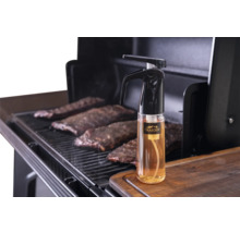 Vaporisateur Traeger pour barbecue sur une planche à découper en bois à côté d'un gril avec des travers de porc.