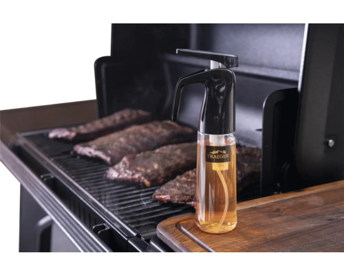 Vaporisateur Traeger pour barbecue sur une planche à découper en bois à côté d'un gril avec des travers de porc.
