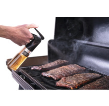 Main tenant un vaporisateur Traeger et vaporisant des travers de porc sur une grille de barbecue alors que de la fumée s'élève.