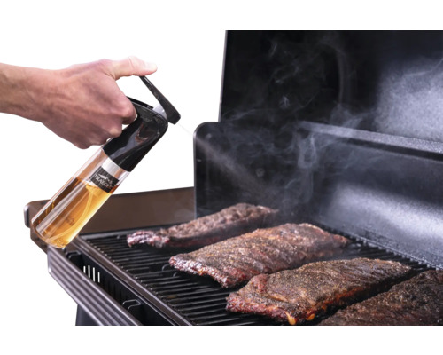 Main tenant un vaporisateur Traeger et vaporisant des travers de porc sur une grille de barbecue alors que de la fumée s'élève.