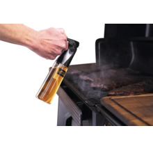 Une main tient un vaporisateur d'huile Traeger au-dessus de la viande en train de cuire sur un barbecue.