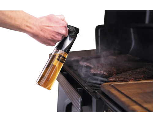 Une main tient un vaporisateur d'huile Traeger au-dessus de la viande en train de cuire sur un barbecue.