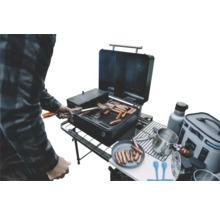 Personne faisant griller des saucisses sur un barbecue portable noir lors d une sortie de camping avec divers accessoires et un sac isotherme.