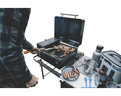 Plancha Traeger Iron Griddle Personne faisant griller des saucisses sur un barbecue portable noir lors d une sortie de camping avec divers accessoires et un sac isotherme.