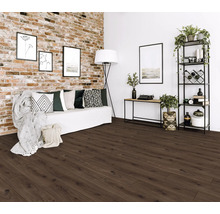 Hell gestaltetes Wohnzimmer mit dunkelbraunem Boden in Holzoptik, einer Klinkerwand und einem weißen Sofa mit gemusterten Kissen.