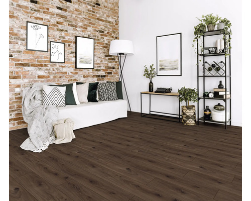 Hell gestaltetes Wohnzimmer mit dunkelbraunem Boden in Holzoptik, einer Klinkerwand und einem weißen Sofa mit gemusterten Kissen.