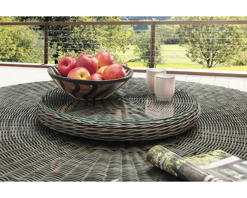 Table de jardin ronde en résine tressée grise avec plateau en verre, avec une coupe de pommes et des tasses sur une terrasse avec vue sur la nature.