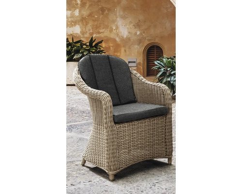 Fauteuil de jardin tressé avec coussins d'assise et de dossier sur une terrasse en pierre devant un mur rustique avec des plantes.