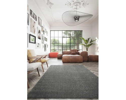 Moderner Wohnraum mit grauem Hochflor-Teppich, braunem Ledersofa, Fischgrät-Parkett und Bilderwand vor großem Fenster.