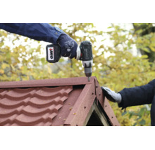 Une personne portant des gants de travail fixe des bardeaux de toiture rouges sur un faîtage avec une perceuse sans fil Metabo.