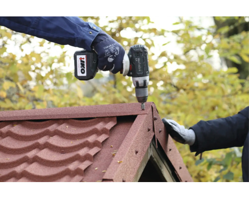 Une personne portant des gants de travail fixe des bardeaux de toiture rouges sur un faîtage avec une perceuse sans fil Metabo.