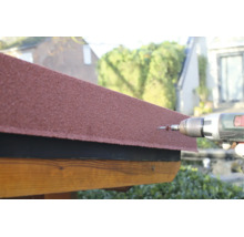 Fixation d'un bardeau bitumé rouge sur une structure en bois avec une perceuse-visseuse sans fil et une vis.