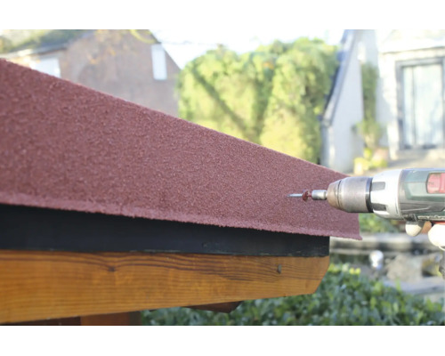 Fixation d'un bardeau bitumé rouge sur une structure en bois avec une perceuse-visseuse sans fil et une vis.
