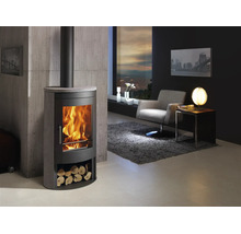 Moderner Kaminofen aus Stahl und Stein mit Feuer und Holzfach in hellem Wohnzimmer mit Sessel, Teppich und Betonoptik-Wand.