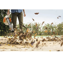 Une personne utilise un souffleur de feuilles pour enlever des feuilles mortes d'une surface pavée dans un jardin.