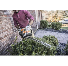 Person schneidet einen Busch mit einer Stihl Benzin-Heckenschere neben einer Ziegelwand im Garten.