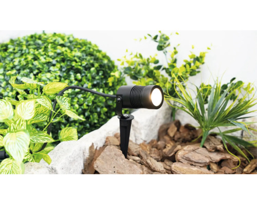 Projecteur de jardin à LED noir avec piquet de terre dans un parterre avec du paillis et des plantes vertes.