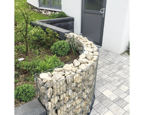 Gebogene Gabione als Beeteinfassung im Garten, gefüllt mit hellen Natursteinen neben einem gepflasterten Weg.