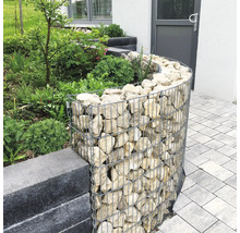 Geschwungene Gabione aus Drahtgeflecht mit Naturstein-Füllung als Beetumrandung im Garten neben einer Pflasterfläche.