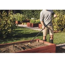 Eine Person benutzt eine Gartenharke, um die Erde in einem hölzernen Hochbeet in einem hellen Garten zu ebnen.