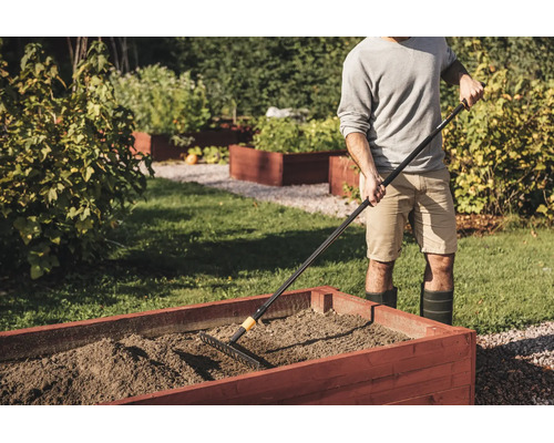 Eine Person benutzt eine Gartenharke, um die Erde in einem hölzernen Hochbeet in einem hellen Garten zu ebnen.