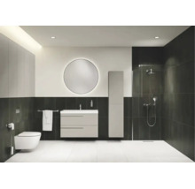 Modernes Badezimmer mit Waschtischunterschrank, Hochschrank, rundem beleuchtetem Spiegel, Wand-Toilette und begehbarer Dusche mit dunklen Fliesen.