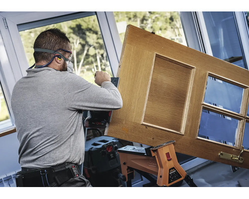Handwerker schleift eine Holztür mit einem Akku-Exzenterschleifer in einer hellen Werkstatt.