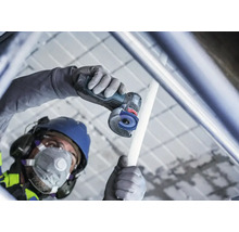Artisan avec casque de protection et masque respiratoire coupe un tuyau avec une meuleuse angulaire sans fil sur un chantier.