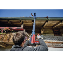 Une personne nettoie une gouttière avec un kit de nettoyage de gouttière sans fil Einhell sur une maison en bois rustique par temps ensoleillé.