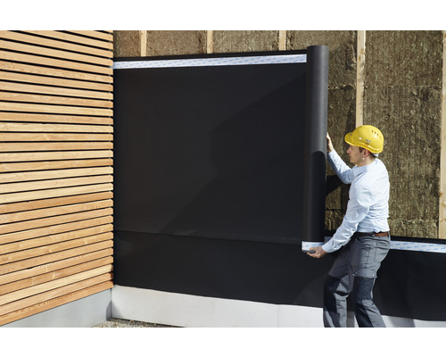 Un homme installe un écran de façade noir sur un mur avec un bardage en bois et une isolation thermique.