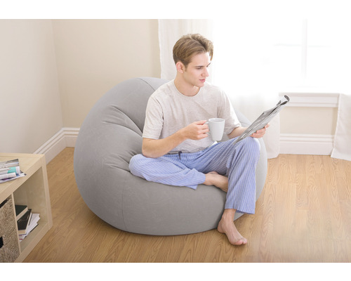 Un homme est assis sur un pouf gris avec un journal et une tasse.