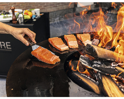 Une personne fait griller du saumon sur une plaque de cuisson avec un feu de bois