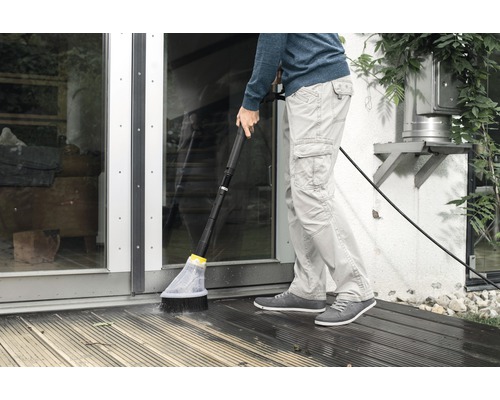 Une personne nettoie une terrasse en bois avec une brosse et un tuyau d''arrosage.