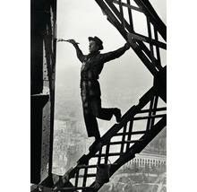 Un homme en tenue de travail et chapeau est en équilibre sur une poutre en acier tout en peignant la structure métallique avec un pinceau.