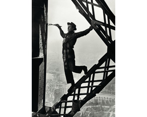 Un homme en tenue de travail et chapeau est en équilibre sur une poutre en acier tout en peignant la structure métallique avec un pinceau.